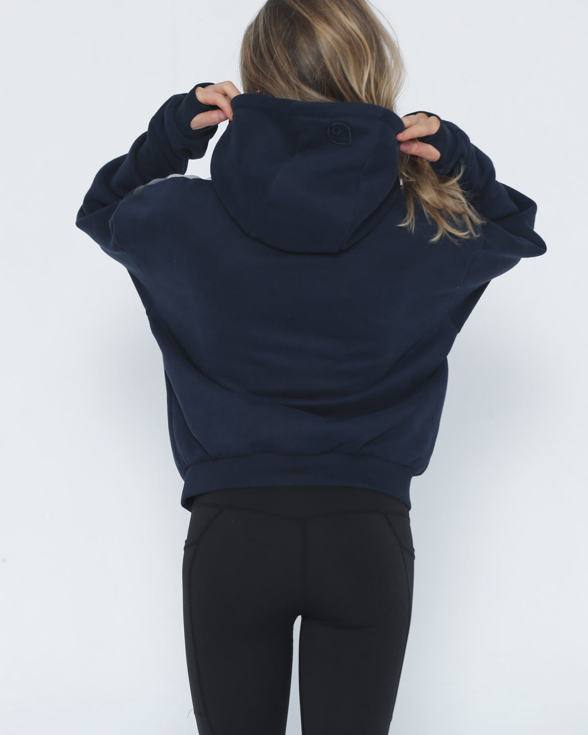 A woman viewed from behind wearing a cropped navy blue hoodie (Ruby) and black leggings, adjusting the hood against a plain white background.