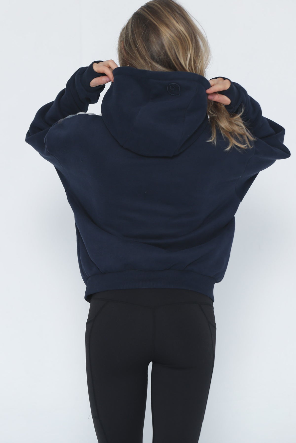 A woman viewed from behind wearing a cropped navy blue hoodie (Ruby) and black leggings, adjusting the hood against a plain white background.