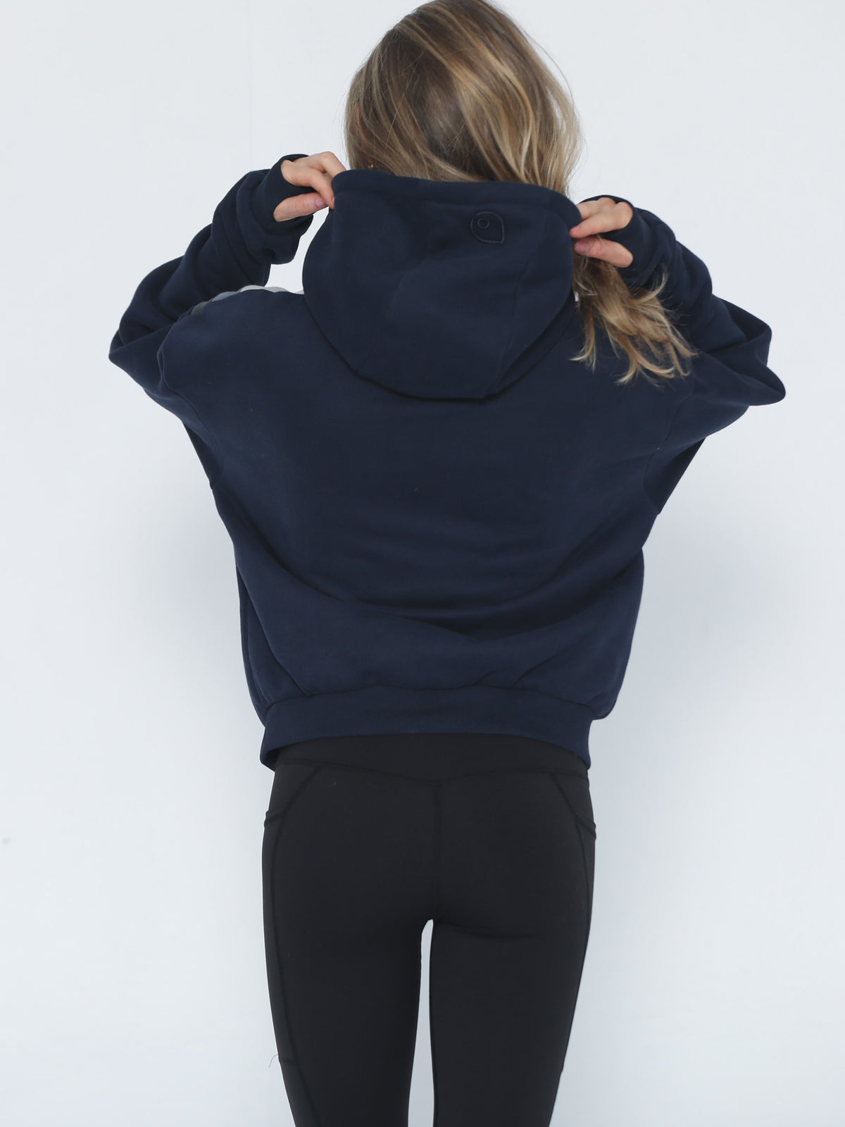 A woman viewed from behind wearing a cropped navy blue hoodie (Ruby) and black leggings, adjusting the hood against a plain white background.
