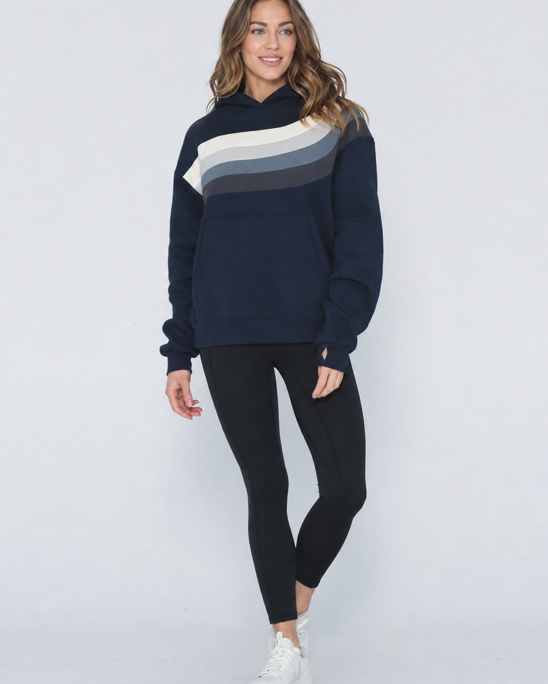 A woman modeling a navy blue sweatshirt with a diagonal color-blocked stripe pattern in cream, light gray, and dark gray across the chest, paired with black leggings and white sneakers against a light gray studio background.