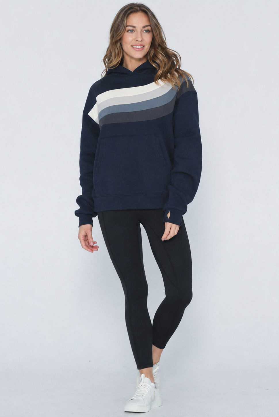 A woman modeling a navy blue sweatshirt with a diagonal color-blocked stripe pattern in cream, light gray, and dark gray across the chest, paired with black leggings and white sneakers against a light gray studio background.