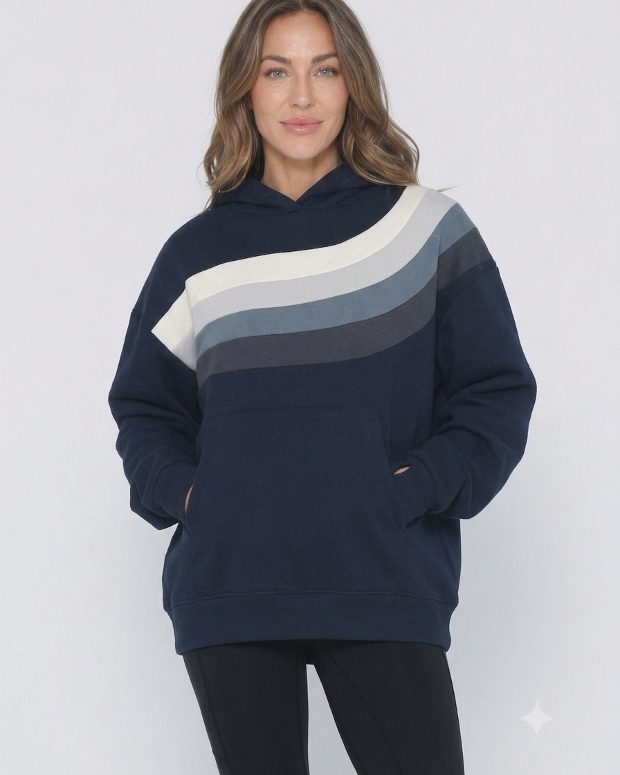 A woman wearing a navy blue hoodie (Ruby) with a diagonal color-blocked stripe pattern in cream, light gray, and medium gray across the chest, paired with black pants, against a light gray studio background.