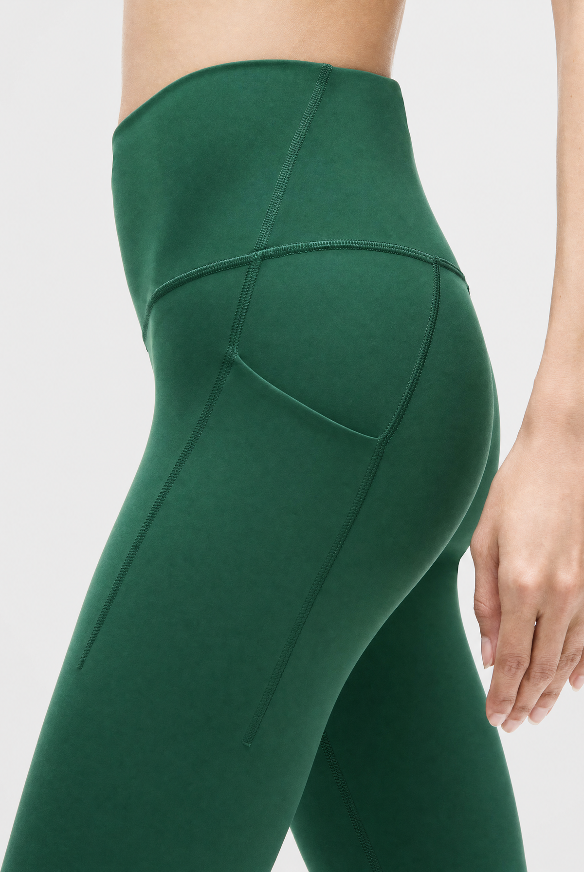 Close-up of a model wearing high-waisted pine green leggings, showing the side pocket detail and seam construction against a light gray background.