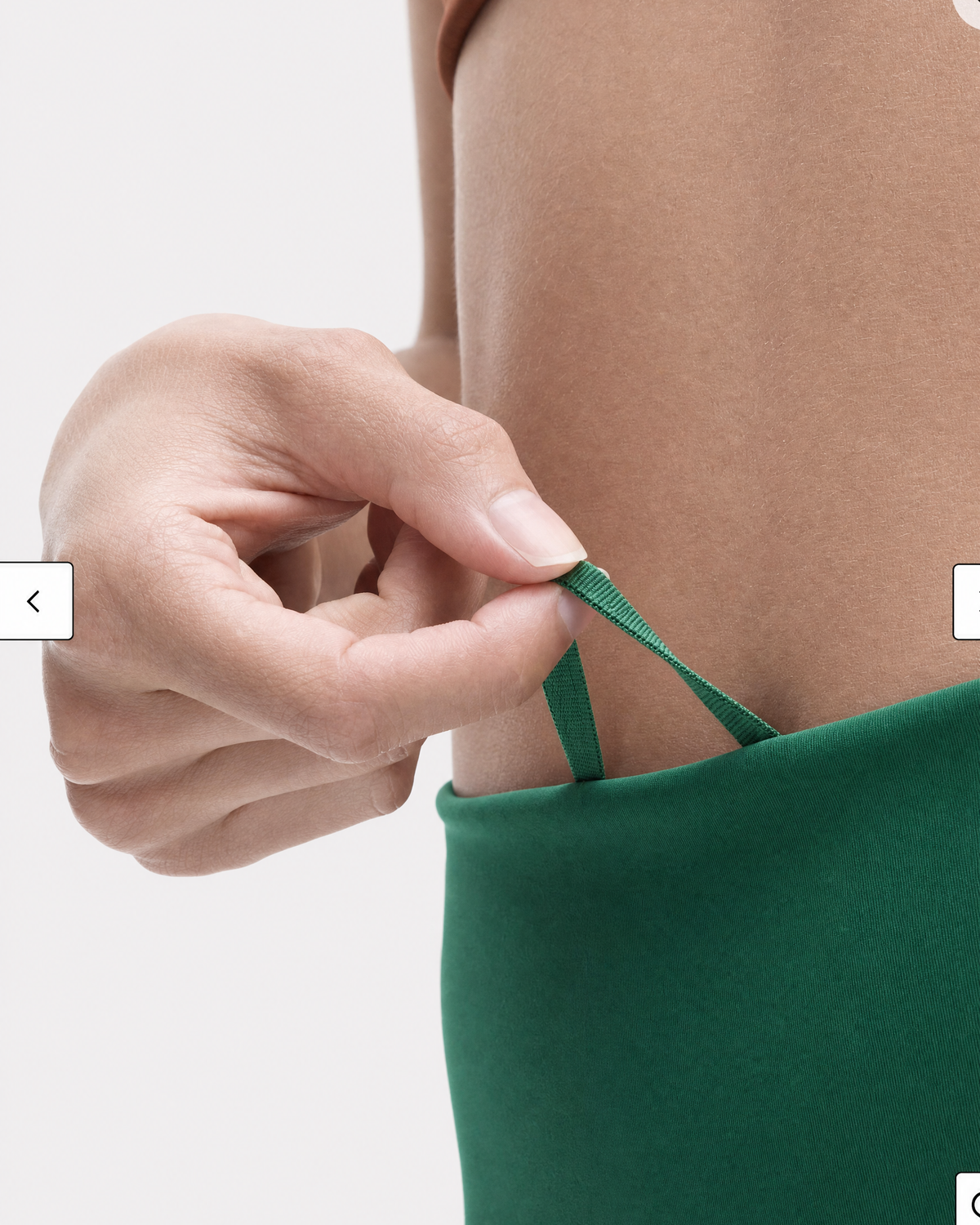 A close-up of a person's hand adjusting a green waist strap on a pine green legging's waist band, shown against a plain light gray background.