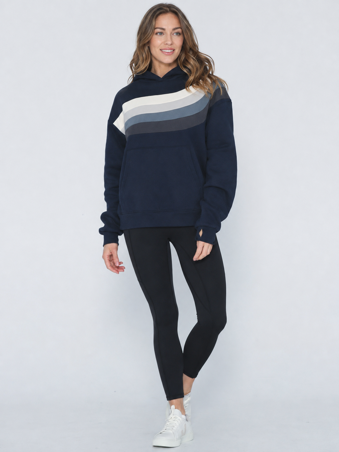A woman modeling a navy blue sweatshirt with a diagonal color-blocked stripe pattern in cream, light gray, and dark gray across the chest, paired with black leggings and white sneakers against a light gray studio background.