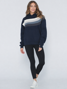 A woman modeling a navy blue sweatshirt with a diagonal color-blocked stripe pattern in cream, light gray, and dark gray across the chest, paired with black leggings and white sneakers against a light gray studio background.
