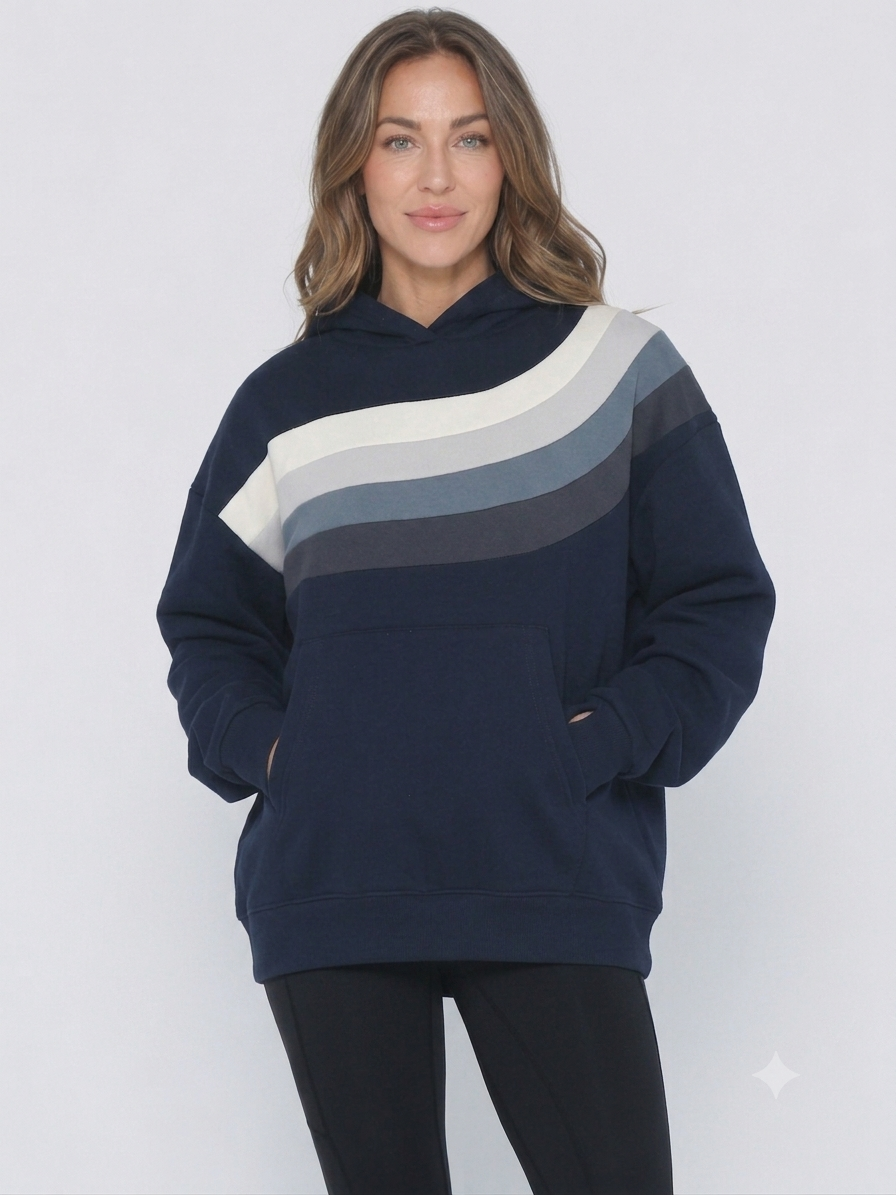 A woman wearing a navy blue hoodie (Ruby) with a diagonal color-blocked stripe pattern in cream, light gray, and medium gray across the chest, paired with black pants, against a light gray studio background.